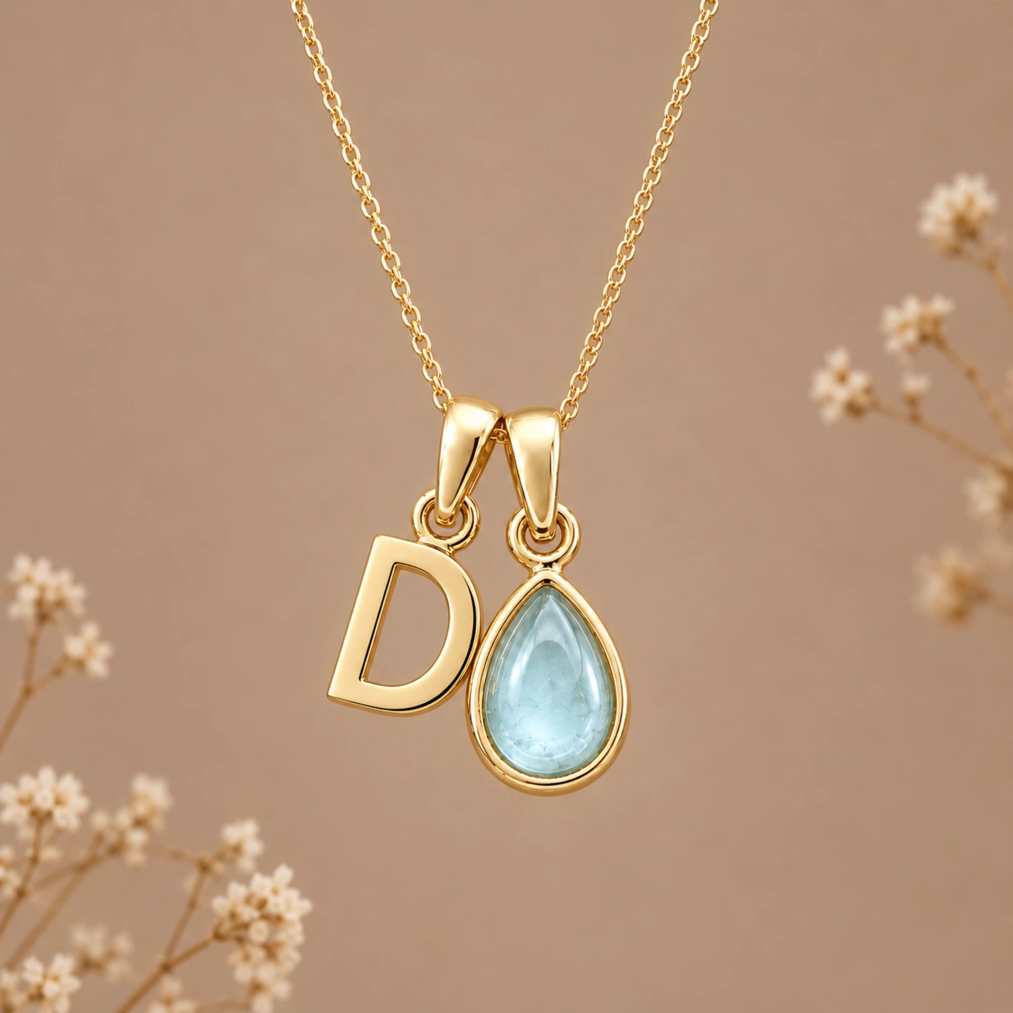 A gold initial march birthstone necklace featuring a letter 'D' pendant and a teardrop-shaped light blue natural aquamarine gemstone pendant on a delicate gold chain, set against a warm beige background with subtle dried flowers.