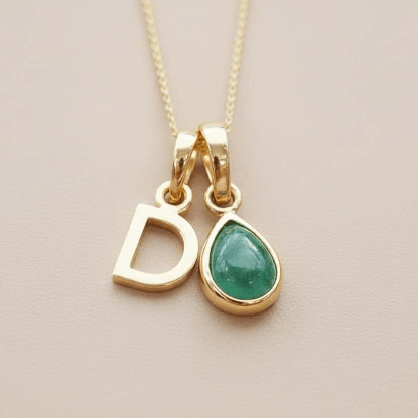 Gold necklace with 'D' pendant and green teardrop charm on a beige background