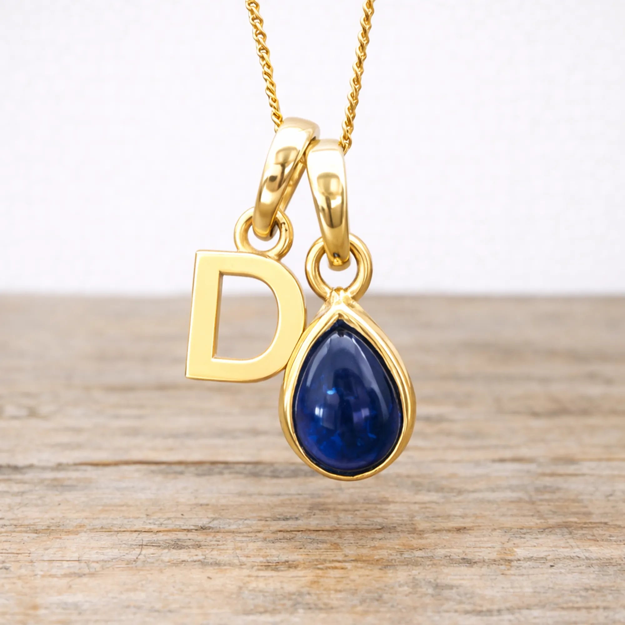 Gold necklace with 'D' pendant and blue sapphire teardrop gemstone on a wooden surface