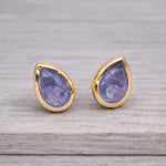 Teardrop-shaped tanzanite gemstone stud earrings in gold settings on a wooden surface