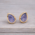 Teardrop-shaped tanzanite gemstone stud earrings in gold settings on a wooden surface