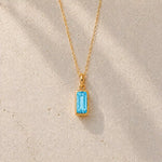 Gold fine chain necklace with a rectangle faceted bezel set december cubic zirconia pendant on a light stone background.
