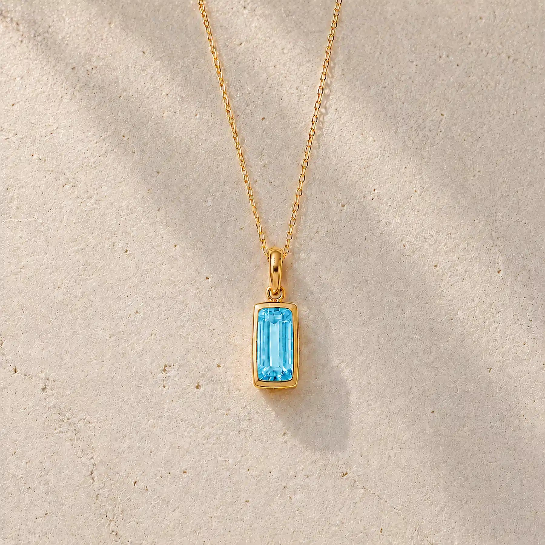 Gold fine chain necklace with a rectangle faceted bezel set december cubic zirconia pendant on a light stone background.