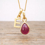 Gold necklace with a teardrop-shaped ruby pendant and an 'E' initial charm on a wooden surface.