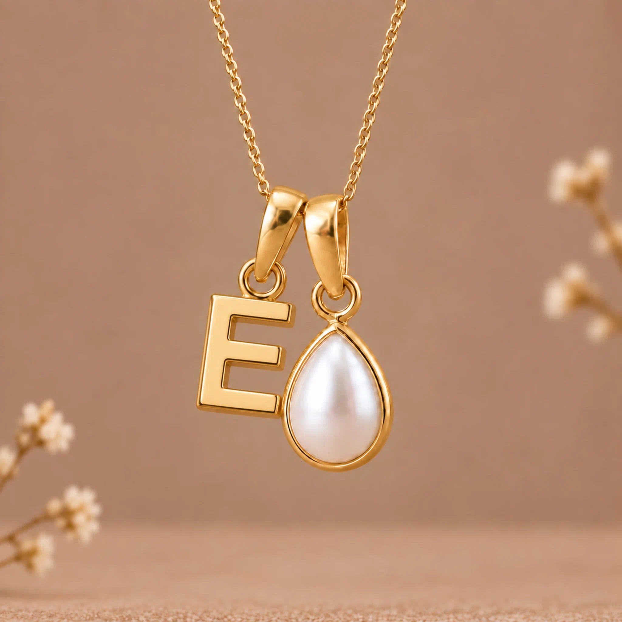 Gold initial pendant necklace featuring a letter 'E' and a teardrop-shaped white pearl on a delicate gold chain, set against a warm beige background with subtle dried flower accents.