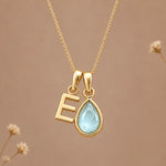 A gold initial march birthstone necklace featuring a letter 'E' pendant and a teardrop-shaped light blue natural aquamarine gemstone pendant on a delicate gold chain, set against a warm beige background with subtle dried flowers.