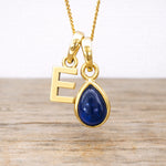 Gold necklace with a blue sapphire teardrop pendant and letter 'E' on a wooden surface.