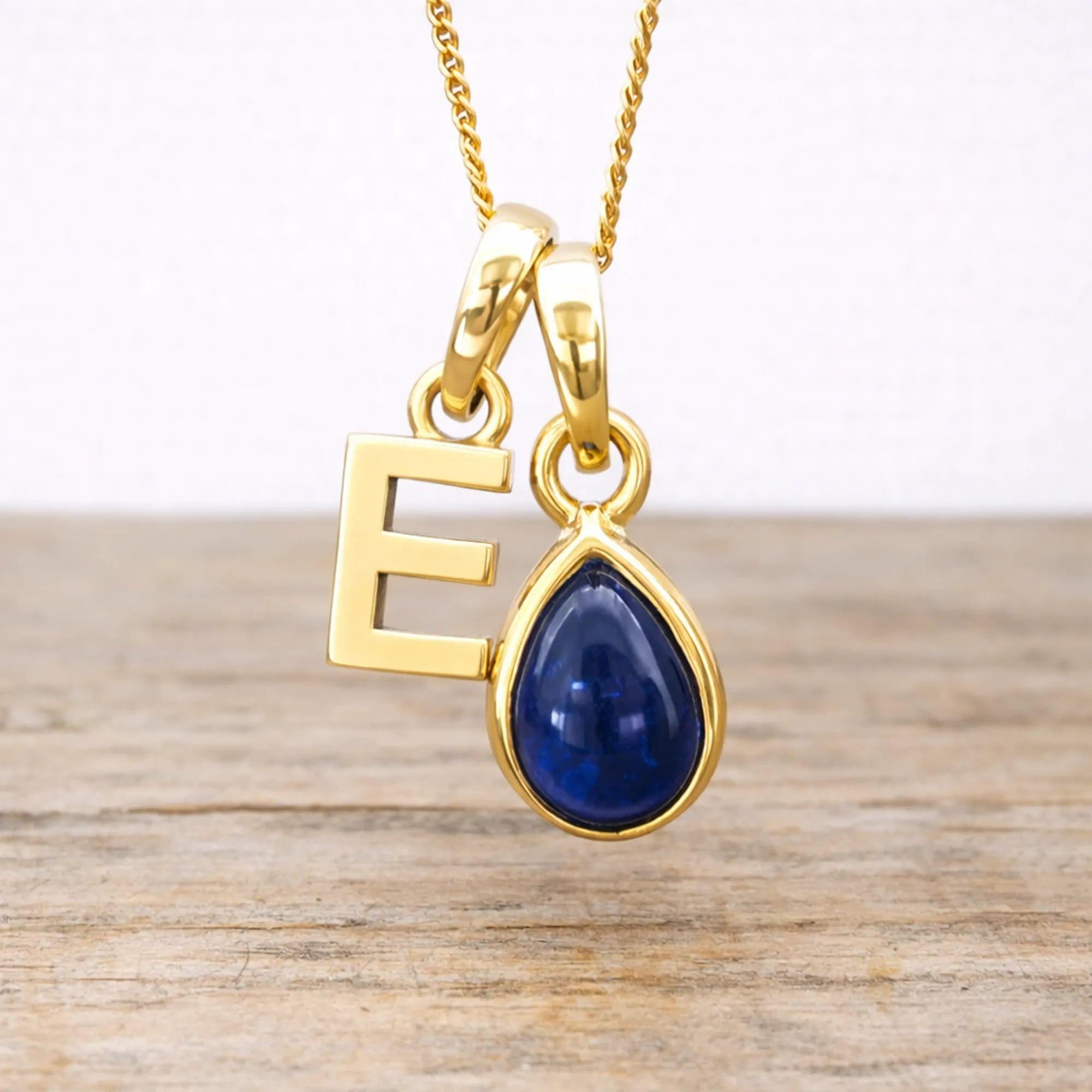 Gold necklace with a blue sapphire teardrop pendant and letter 'E' on a wooden surface.