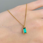 Gold necklace with a green pendant on a hand against a gray background