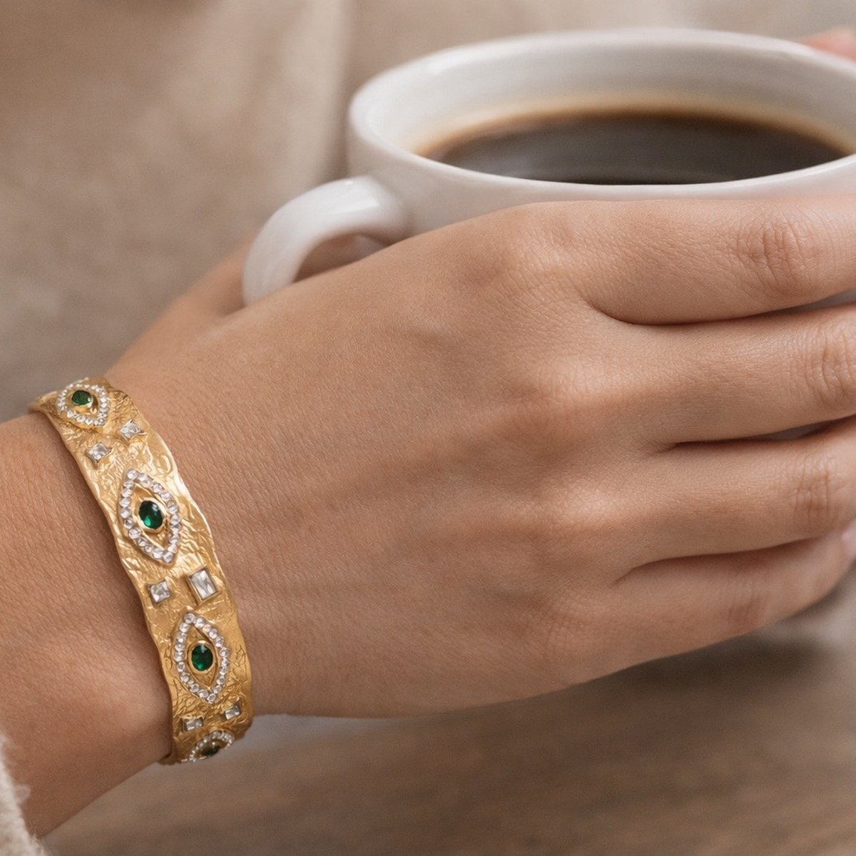 Hand holding a white mug of coffee with a gold bracelet featuring green stones.