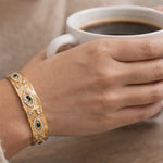 Hand holding a white mug of coffee with a gold bracelet featuring green stones.