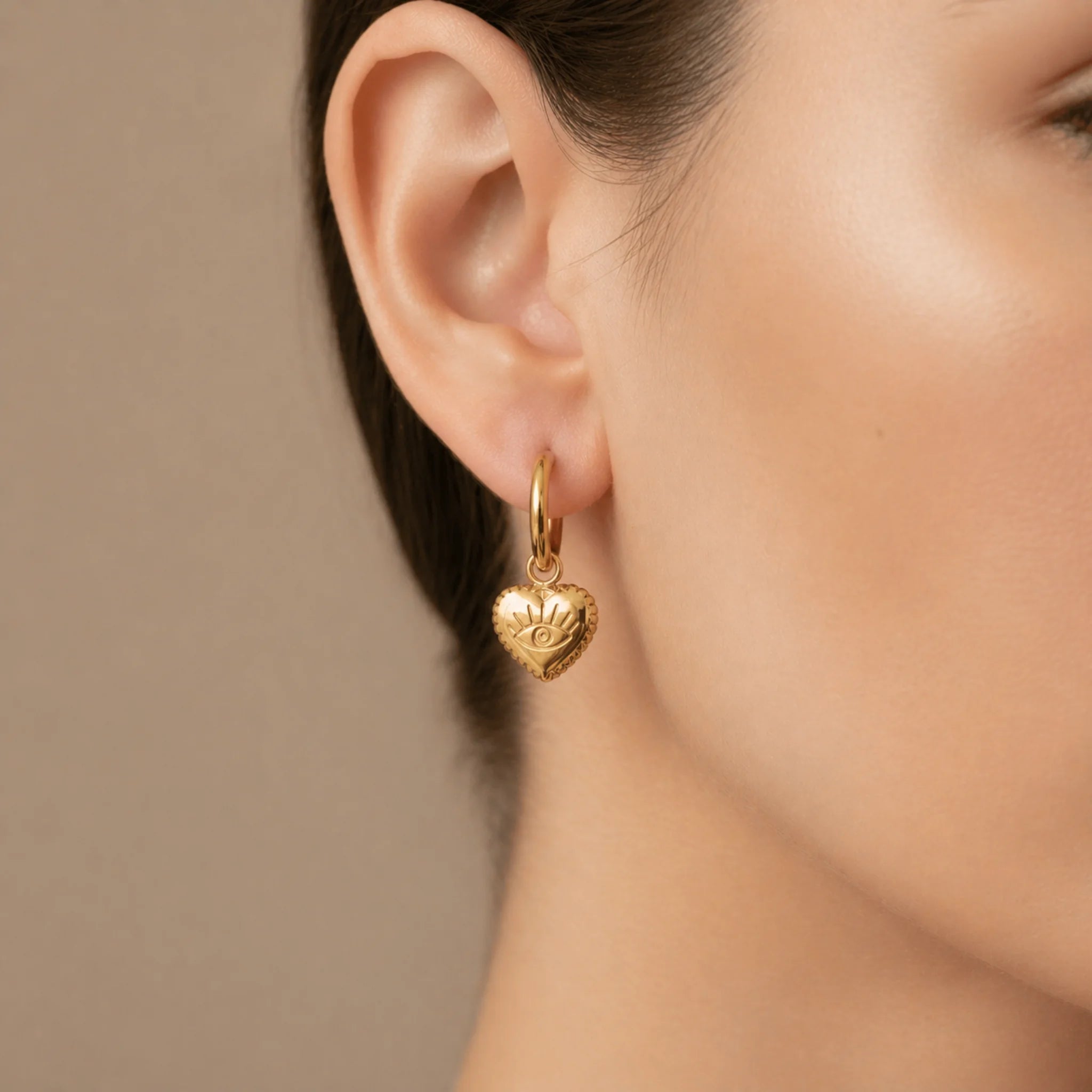 stainless steel waterproof Gold evil eye heart charm huggie earrings worn by a person with a neutral background