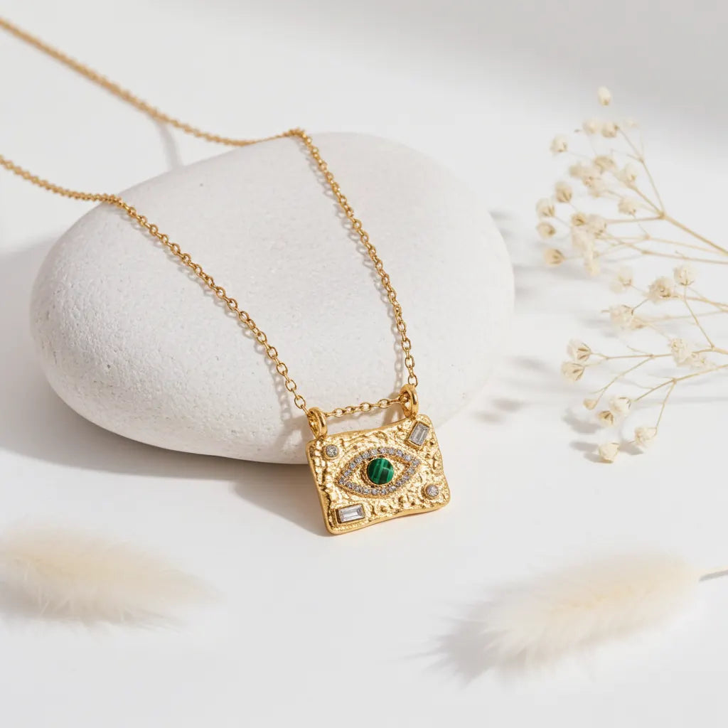 Gold Evil Eye pendant necklace with green malachite stone styled against a white pebble and dried flowers.