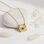 Gold Evil Eye pendant necklace with green malachite stone styled against a white pebble and dried flowers.