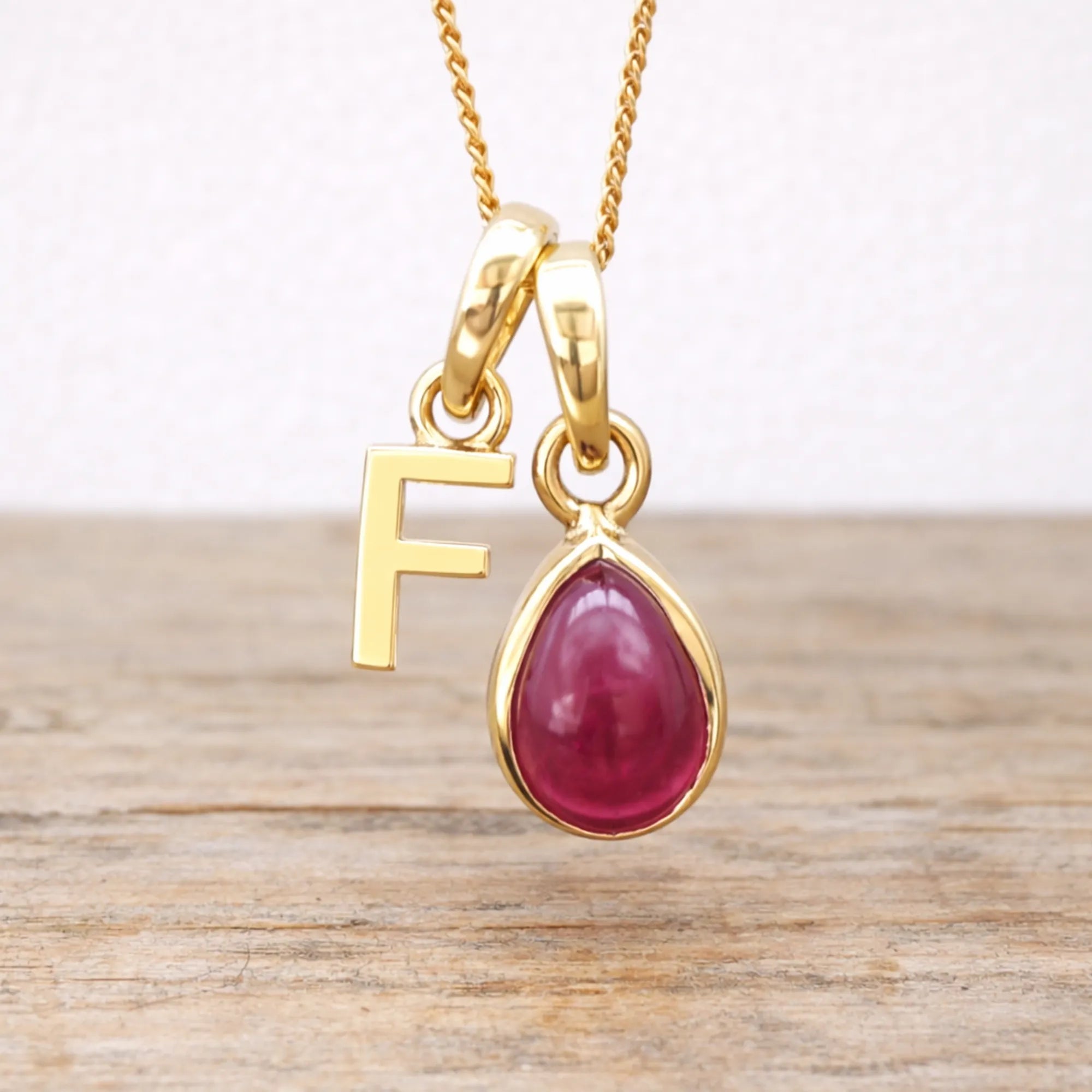 Gold necklace with a red ruby teardrop pendant and letter 'F' charm on a wooden surface.