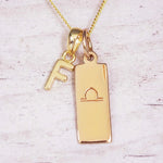 Gold initial and Libra zodiac necklace with a letter F charm and engraved bar pendant on whitewashed wood