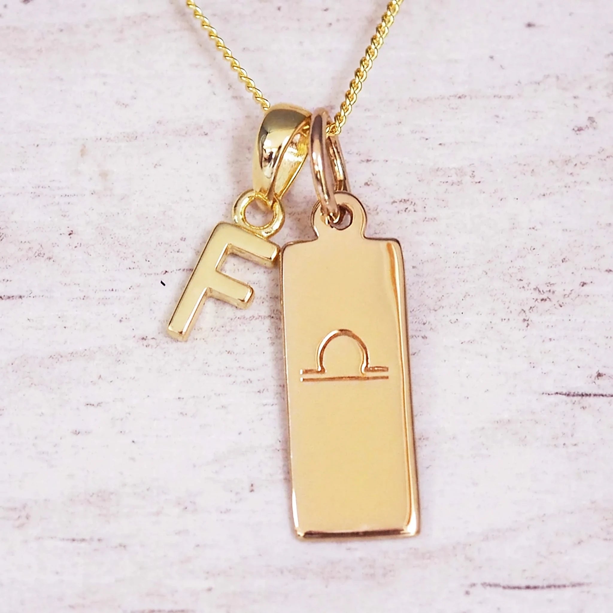 Gold initial and Libra zodiac necklace with a letter F charm and engraved bar pendant on whitewashed wood