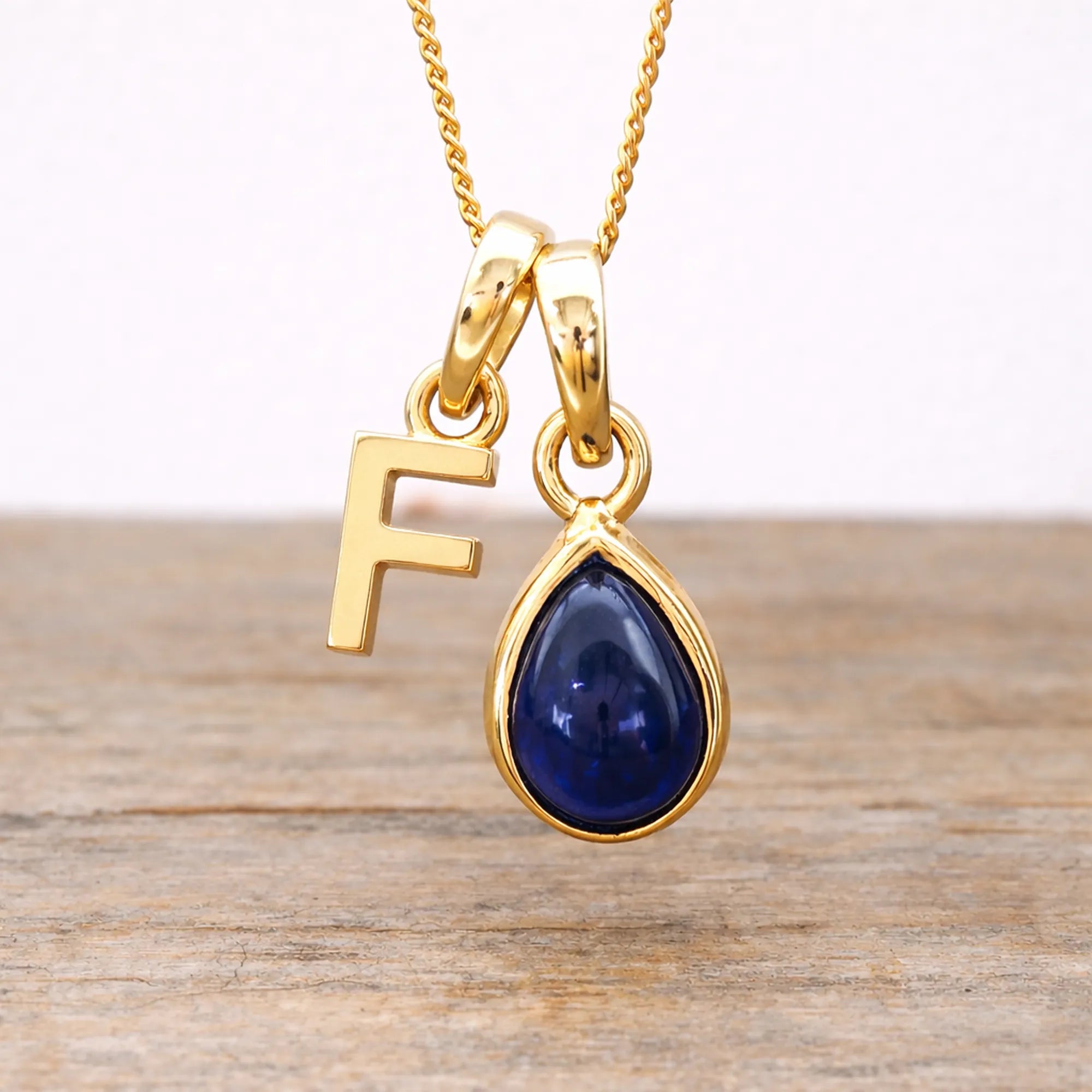 Gold necklace with a blue sapphire teardrop pendant and letter 'F' charm on a wooden surface.
