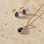 Gold necklace and earrings jewelry set with purple amethyst stones on a beige surface