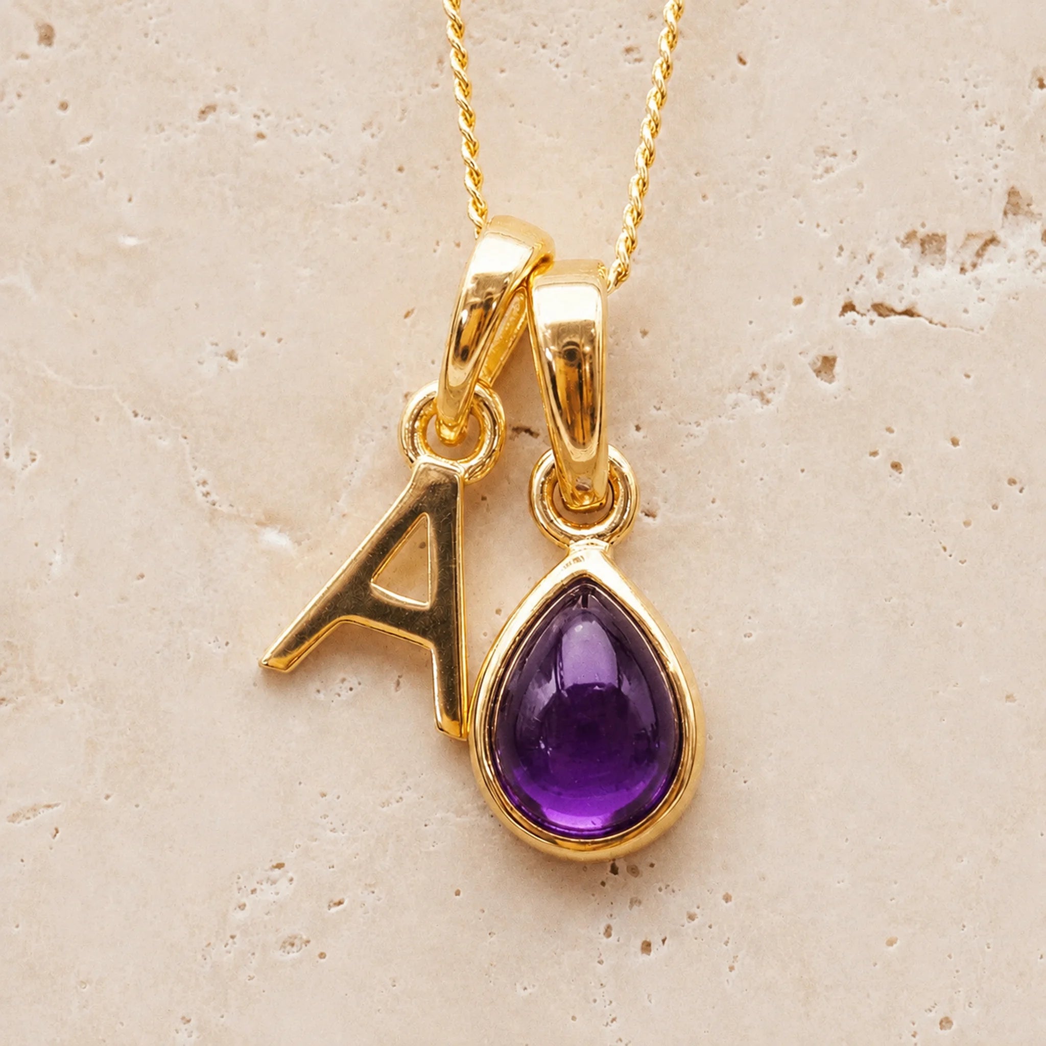 Gold double pendant necklace with a february birthstone purple teardrop amethyst gemstone pendant and letter 'A' charm on a beige background
