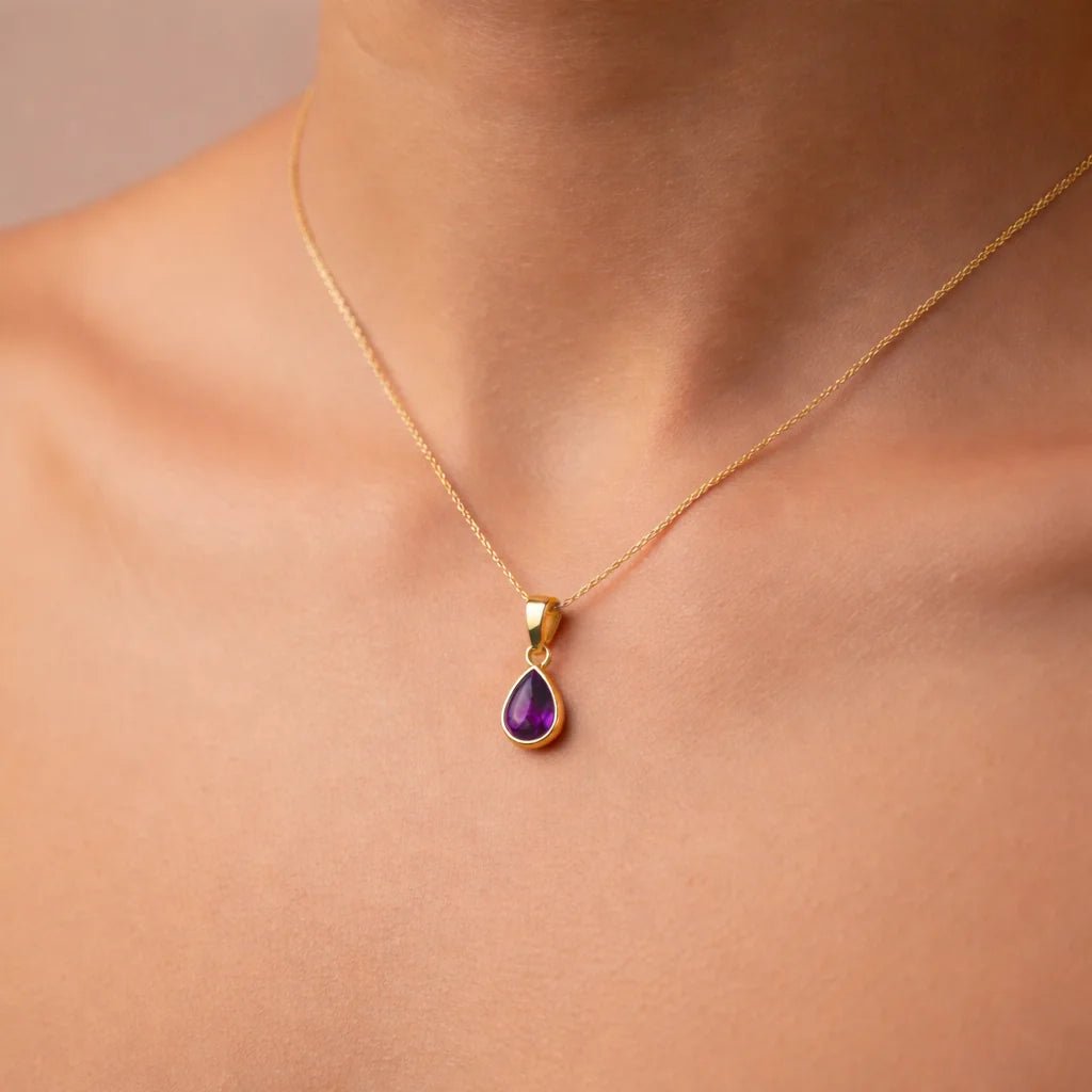 Gold necklace with a purple teardrop natural amethyst gemstone pendant on a person's neck against a neutral background