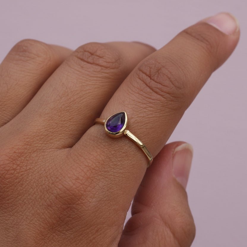 Gold ring with a purple amethyst gemstone on a hand against a pink background