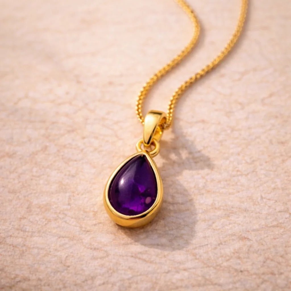 Gold necklace with a purple teardrop natural amethyst gemstone pendant on a beige background