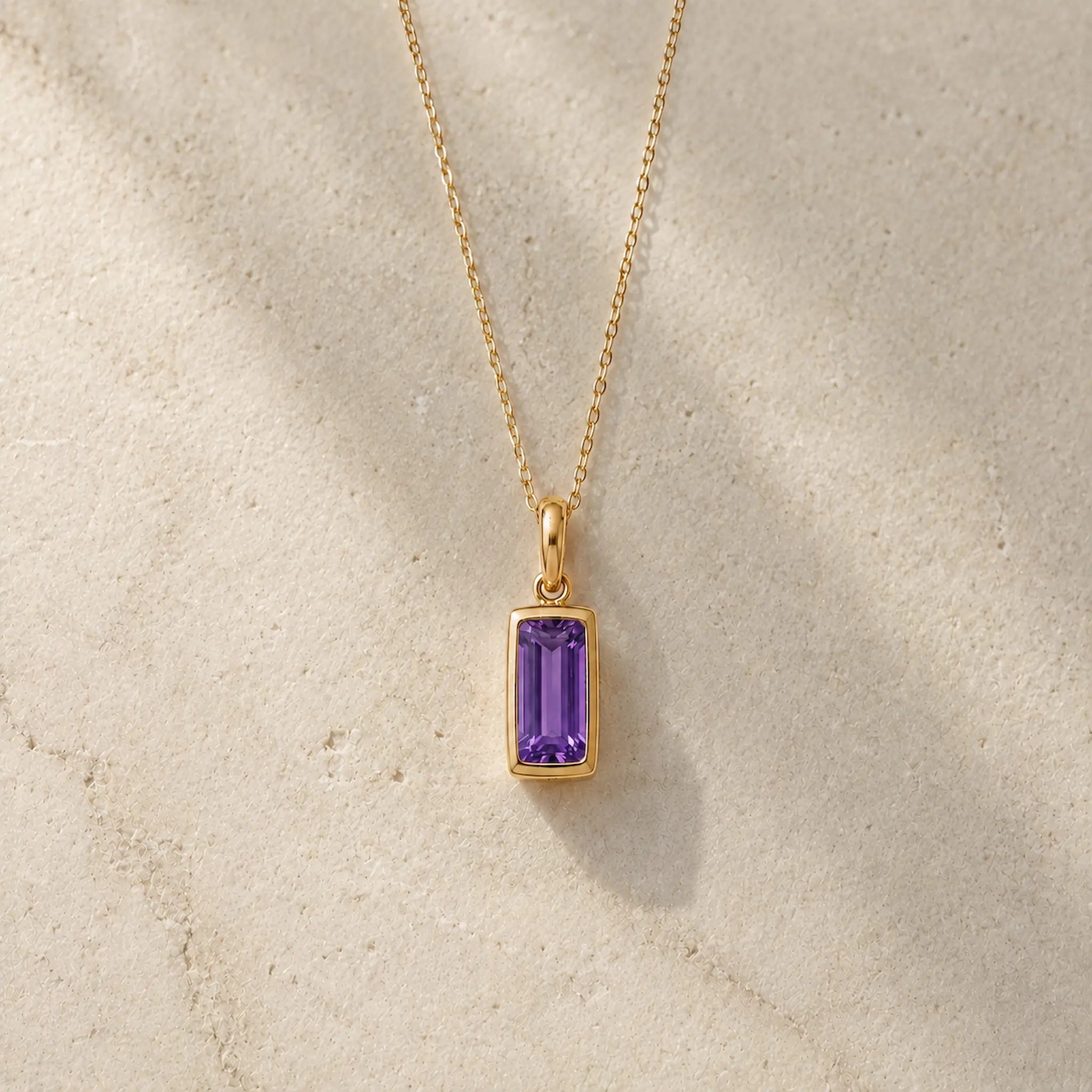 Gold fine chain necklace with a rectangle faceted bezel set february cubic zirconia pendant on a light stone background.