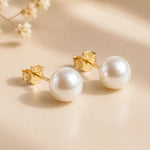 A pair of gold stud pearl earrings with round white freshwater pearls, displayed on a warm beige surface with soft shadows and dried flowers in the background.