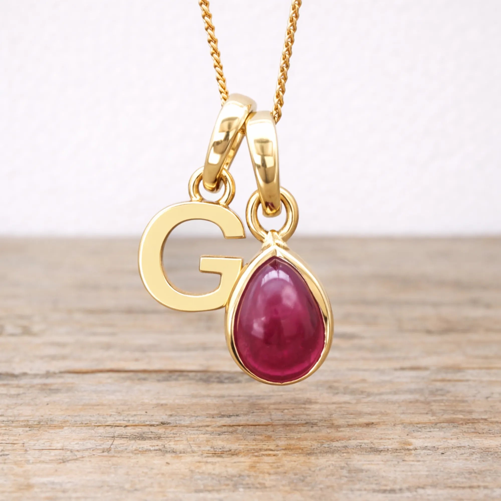 Gold necklace with a red ruby teardrop pendant and letter 'G' charm on a wooden surface.