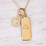 Gold initial and Libra zodiac necklace with a letter G charm and engraved bar pendant on whitewashed wood