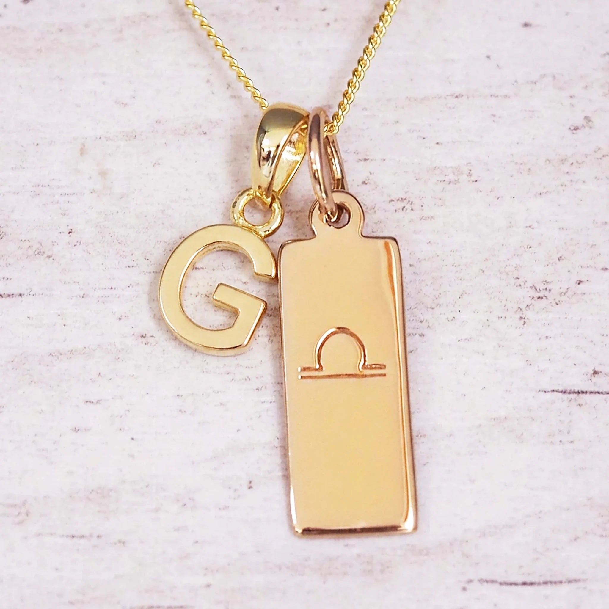 Gold initial and Libra zodiac necklace with a letter G charm and engraved bar pendant on whitewashed wood