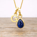 Gold necklace with 'G' pendant and blue sapphire teardrop charm on a wooden surface