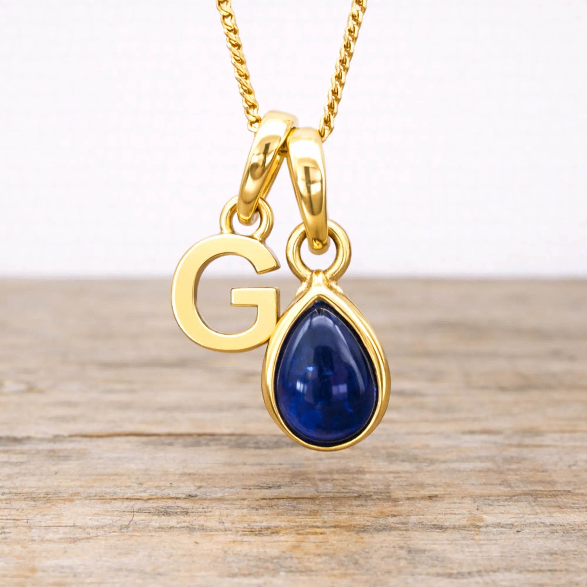 Gold necklace with 'G' pendant and blue sapphire teardrop charm on a wooden surface