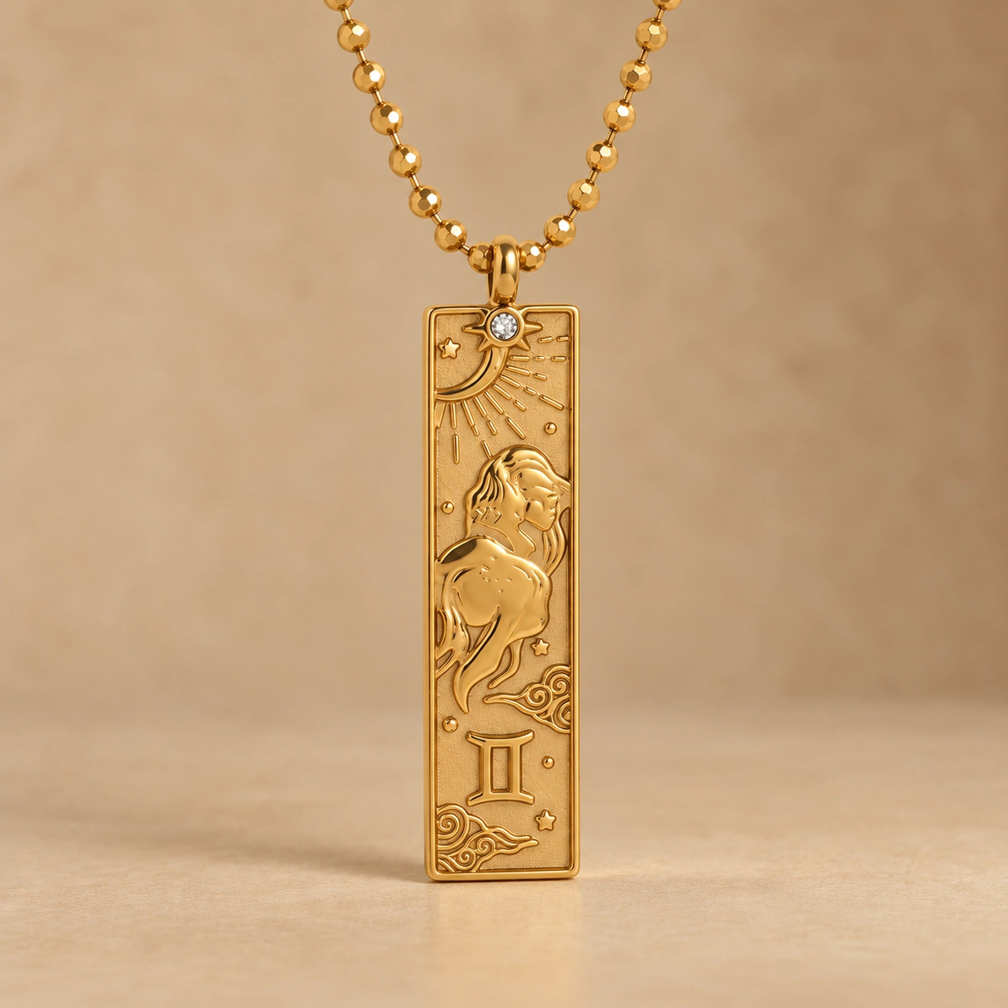 Gold Gemini zodiac necklace, embossed bar pendant with sign symbol on a faceted ball chain, warm beige background