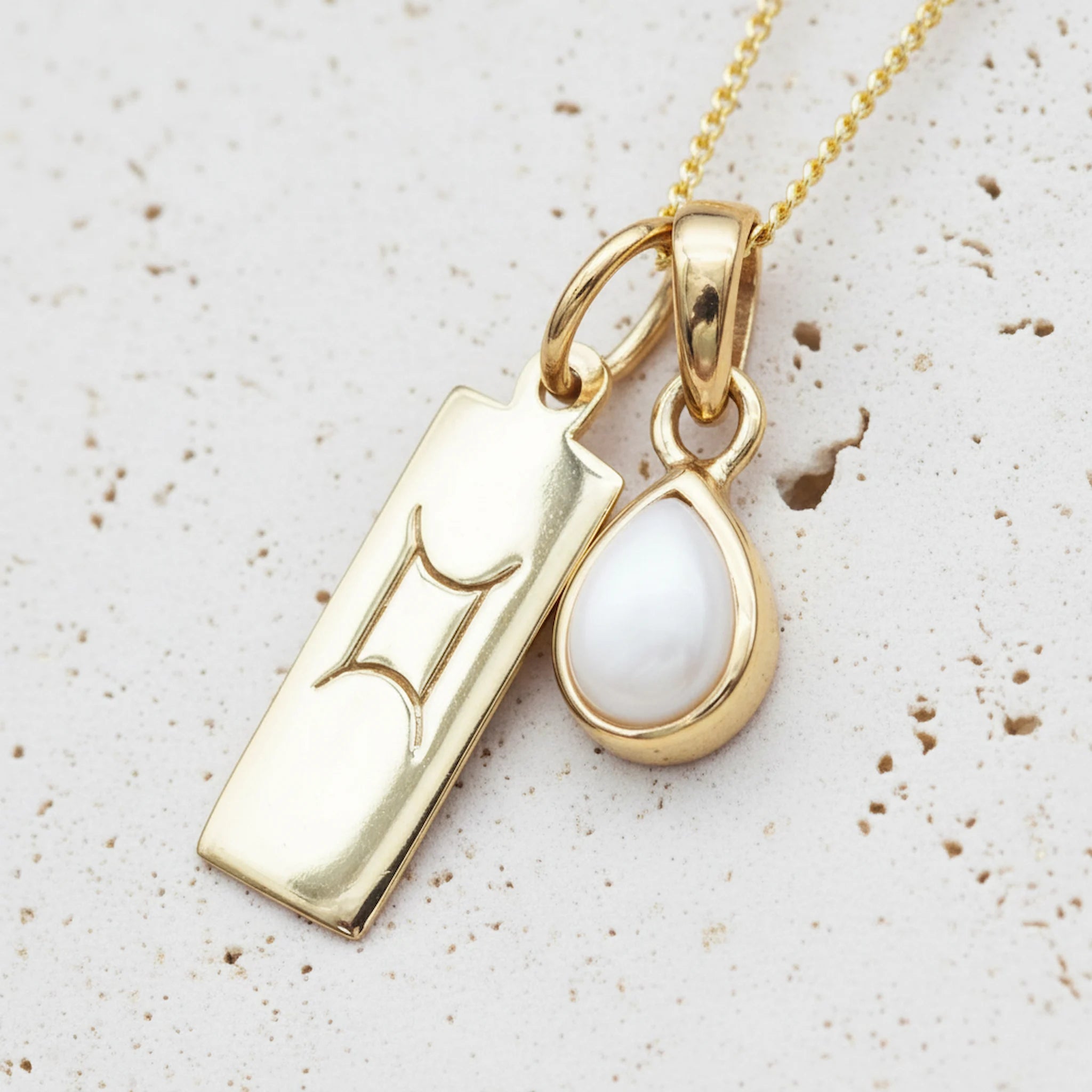 Gold necklace with a tear drop june birthstone pearl pendant with a gemini zodiac pendant on a light background