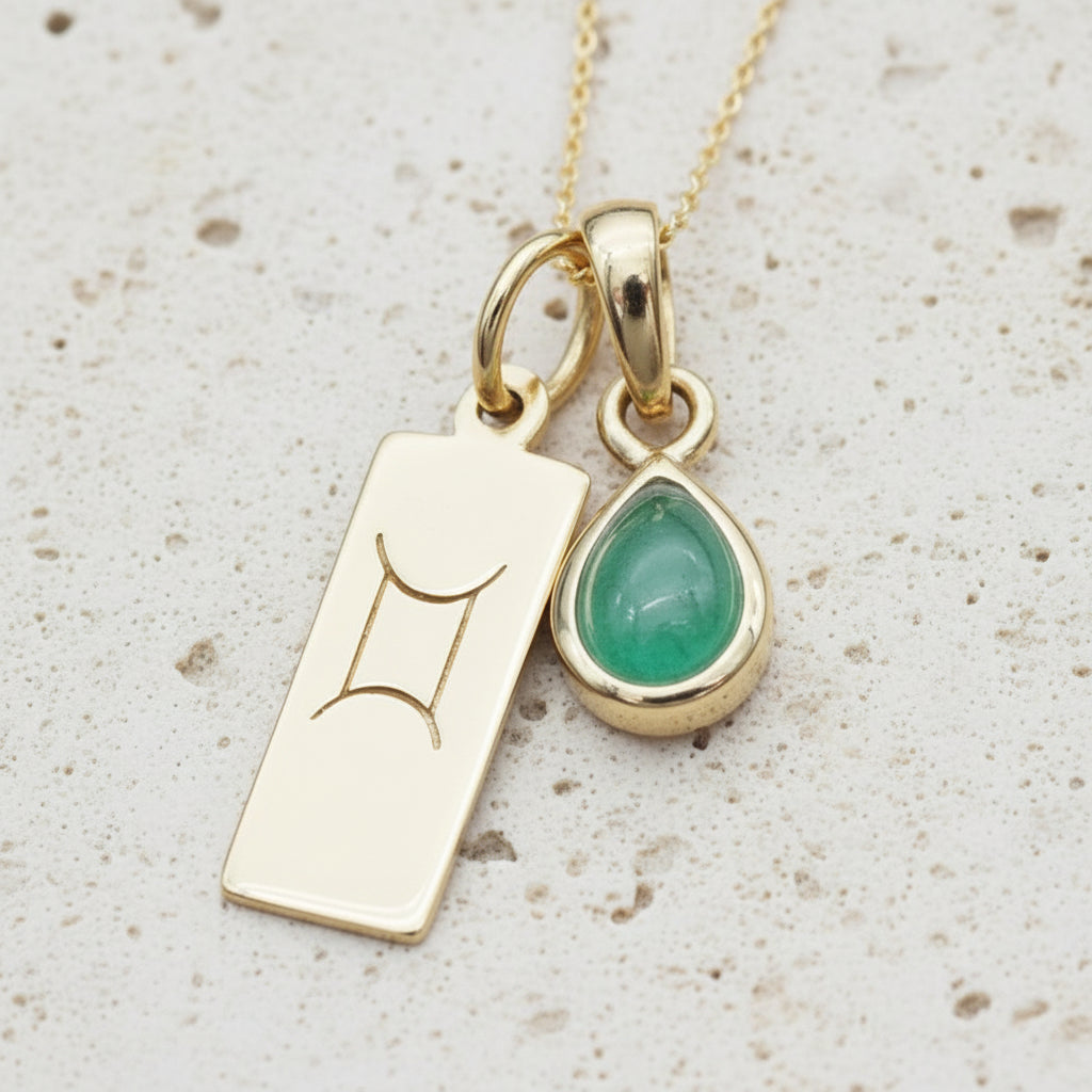 Gold necklace with a green emerald teardrop pendant and a rectangular Gemini zodiac on a wooden surface
