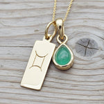 Gold necklace with a green emerald teardrop pendant and a rectangular Gemini zodiac on a wooden surface