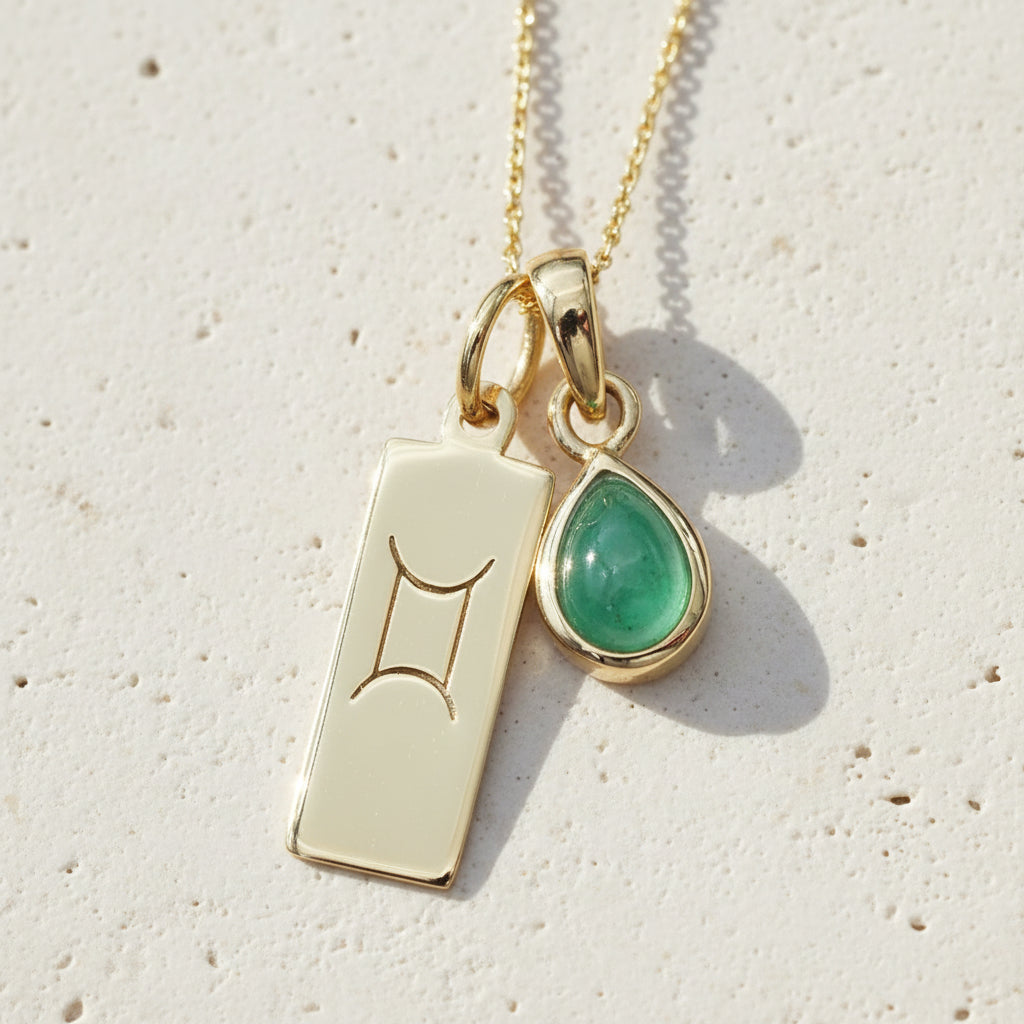 Gold Necklace with Gemini Zodiac symbol pendant and tear drop shape natural Emerald gemstone pendant on a textured surface