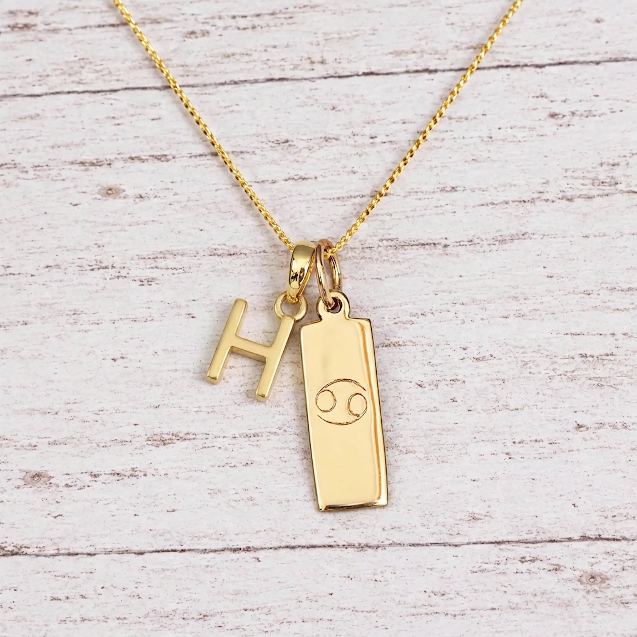 Gold Initial H Pendant and Cancer Zodiac Pendant Necklace on a fine gold chain against a light wooden background