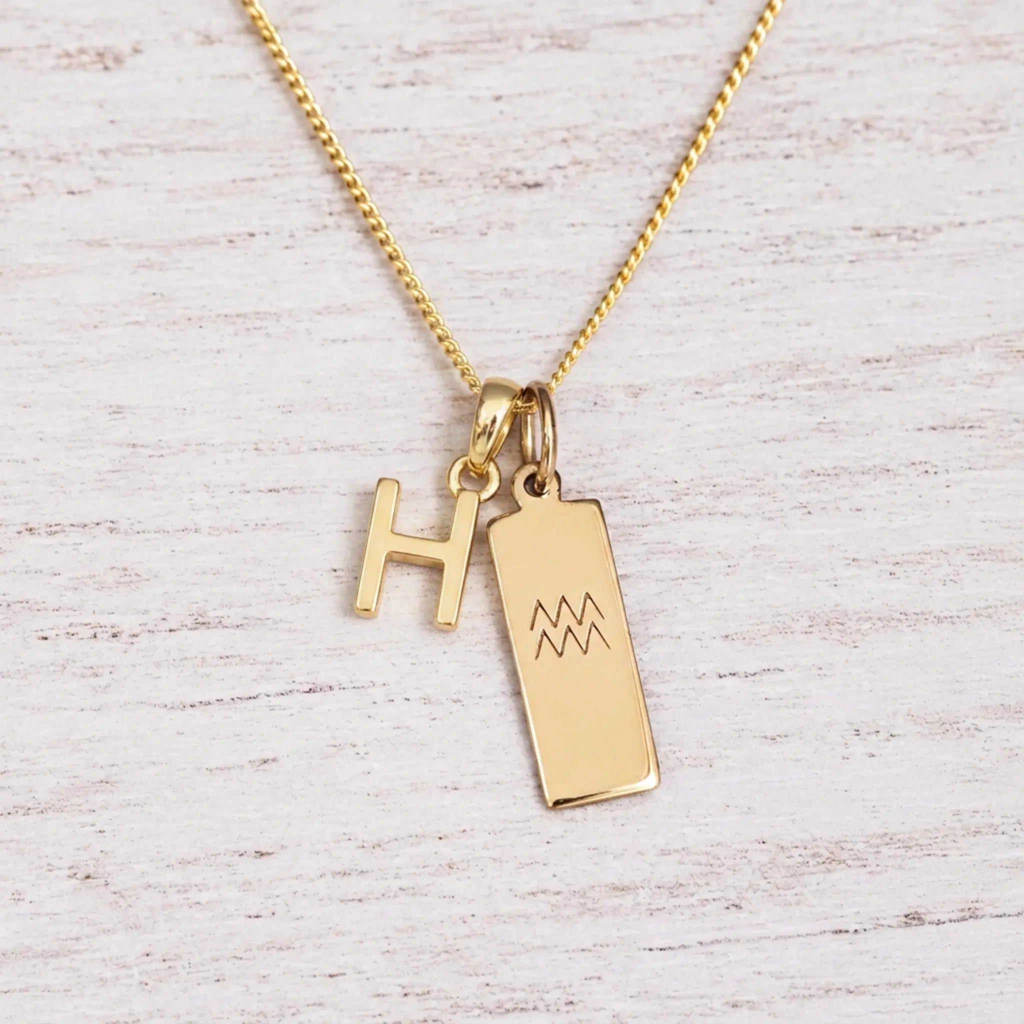 gold H initial and aquarius zodiac necklace on fine gold chain against a light wooden background