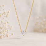A delicate gold chain necklace with a heart-shaped clear gemstone pendant, displayed against a soft beige background with blurred white flowers.