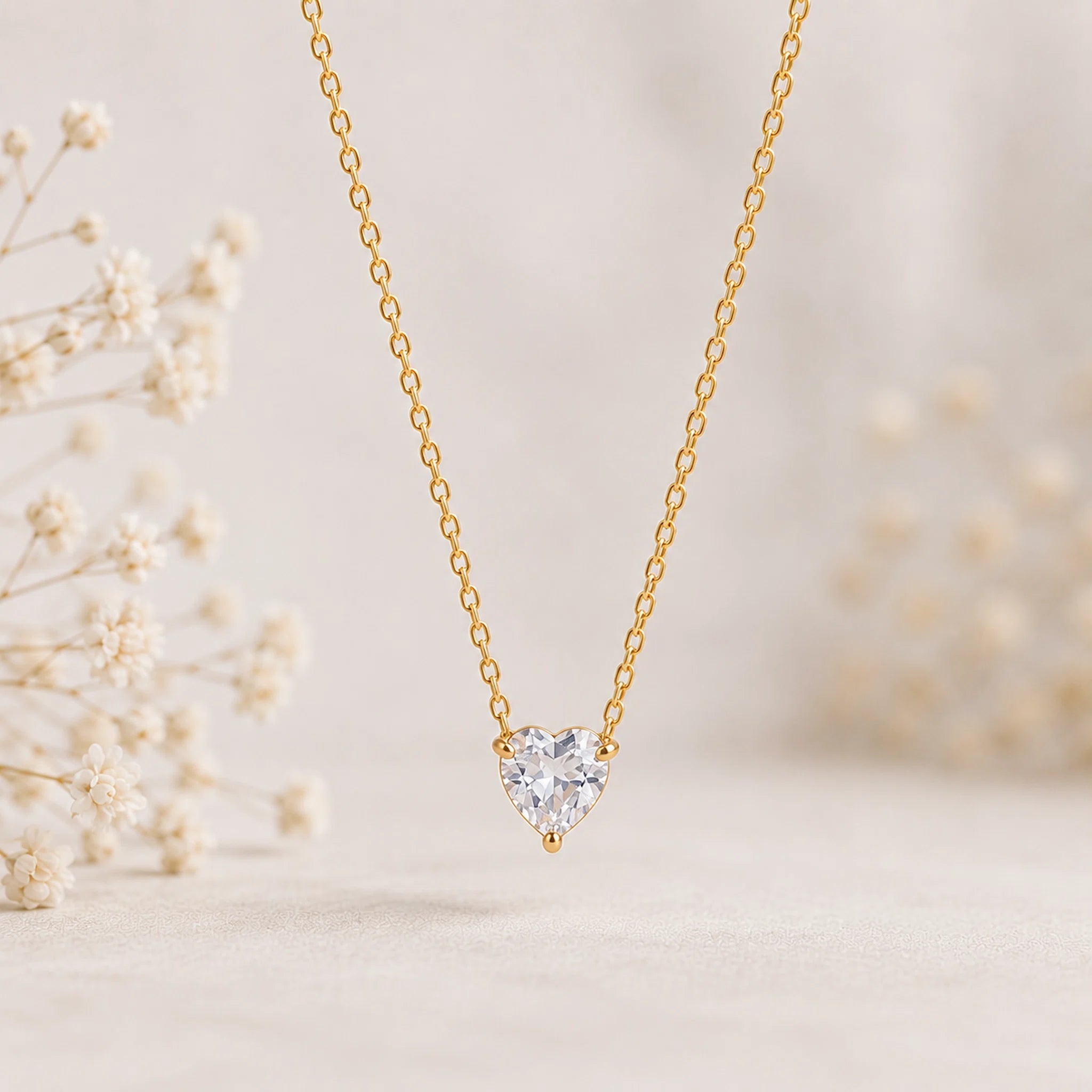 A delicate gold chain necklace with a heart-shaped clear gemstone pendant, displayed against a soft beige background with blurred white flowers.