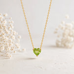 A delicate gold chain necklace with a heart-shaped green gemstone pendant, displayed against a soft, light background with blurred white flowers on either side.