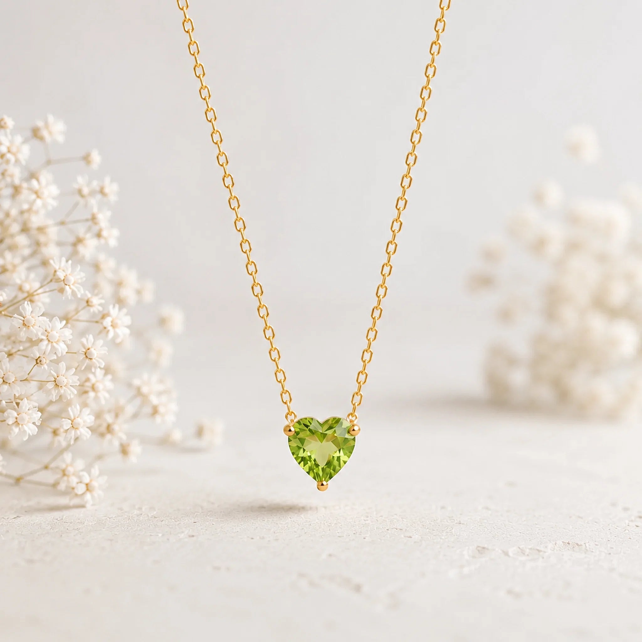A delicate gold chain necklace with a heart-shaped green gemstone pendant, displayed against a soft, light background with blurred white flowers on either side.