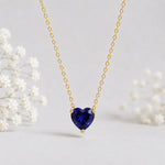 A delicate gold chain necklace with a heart-shaped dark blue gemstone pendant, displayed against a soft light gray background with blurred white baby's breath flowers.