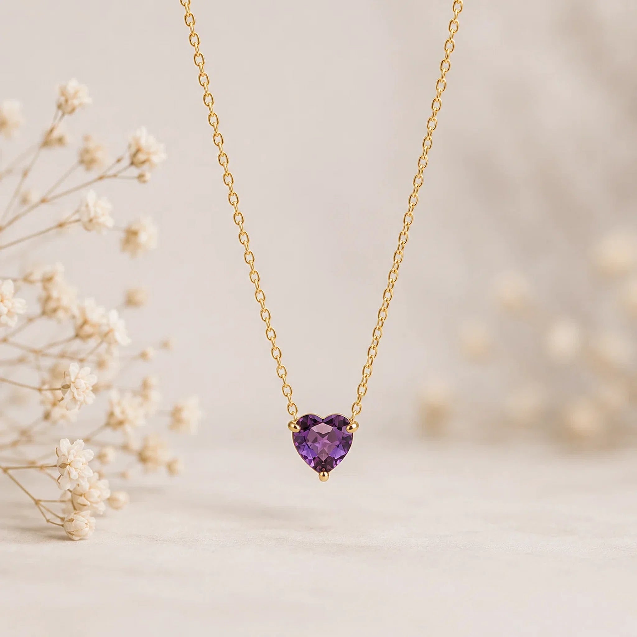 A delicate gold chain necklace with a small heart-shaped purple gemstone pendant, displayed against a soft beige background with dried white flowers on the left.