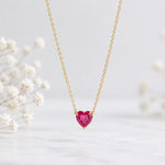 A delicate gold chain necklace with a heart-shaped pink gemstone pendant, displayed against a soft white background with blurred white flowers.