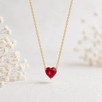 A delicate gold chain necklace featuring a heart-shaped red gemstone pendant, displayed against a soft, light background with blurred white flowers on the sides.