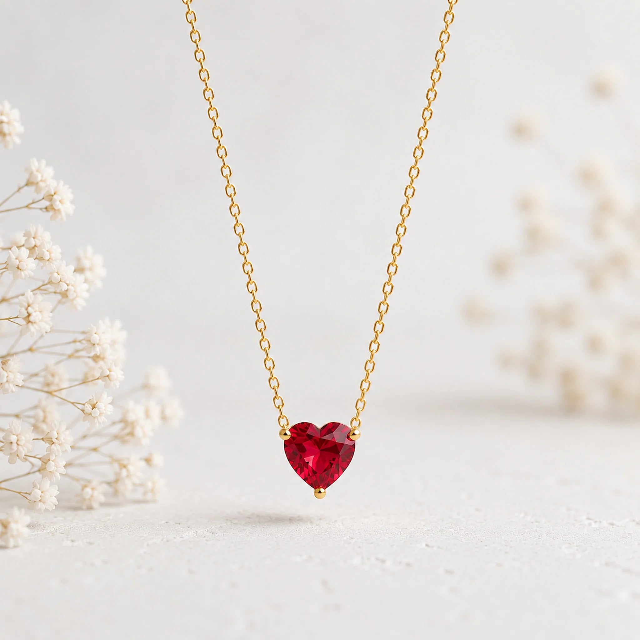 A delicate gold chain necklace featuring a heart-shaped red gemstone pendant, displayed against a soft, light background with blurred white flowers on the sides.